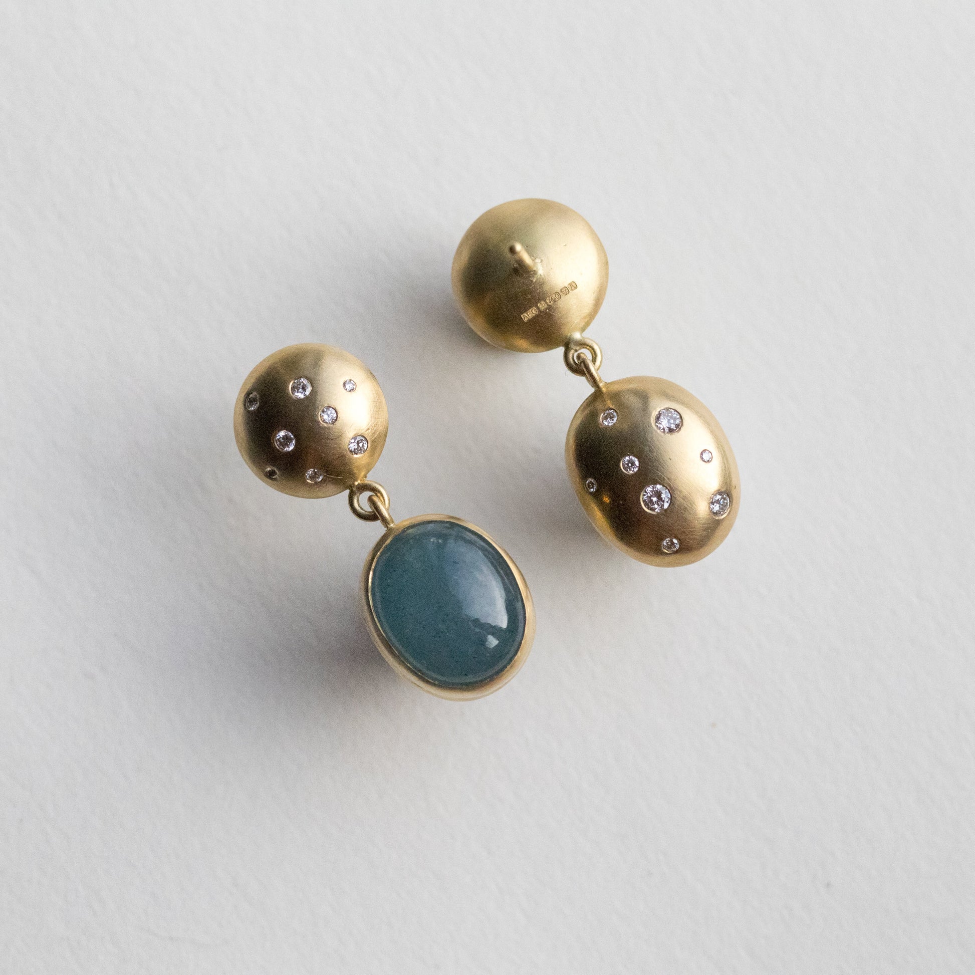 Statement 18ct yellow gold diamond set earrings featuring an oval blue cabochon aquamarine gemstone in a smooth bezel setting, back speckled with diamonds paper shots angle