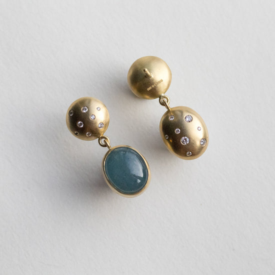 Statement 18ct yellow gold diamond set earrings featuring an oval blue cabochon aquamarine gemstone in a smooth bezel setting, back speckled with diamonds paper shots angle