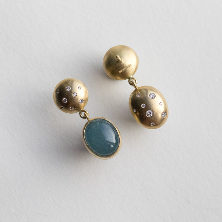Statement 18ct yellow gold diamond set earrings featuring an oval blue cabochon aquamarine gemstone in a smooth bezel setting, back speckled with diamonds paper shots angle