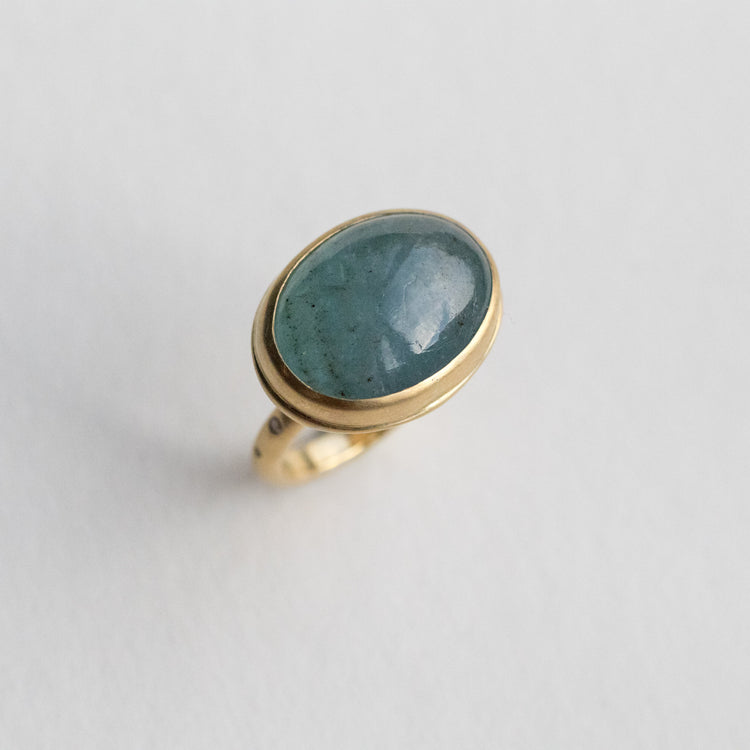 Angled view of an 18ct yellow gold ring with an oval aquamarine blue cabochon stone, highlighting diamond accents on the band