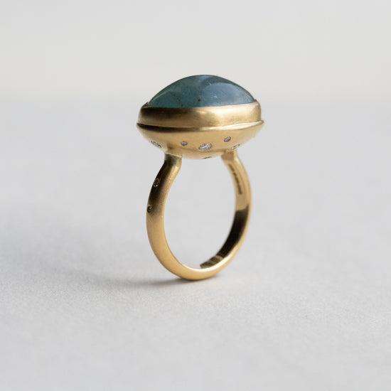 Angled view of an 18ct yellow gold ring with an oval aquamarine blue cabochon stone, highlighting diamond accents on the band and underneath the setting 