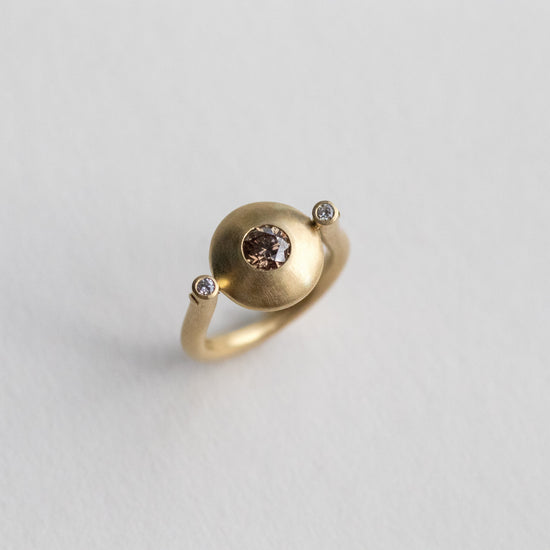 Modern 18ct yellow gold ring with a matte circular spinning disc speckle set with a central natural brown cognac diamond and white side diamonds, paper shot at angle