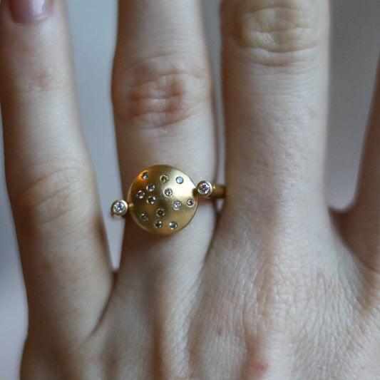 Modern 18ct yellow gold ring with a matte circular spinning disc speckle set with a central natural brown cognac diamonds and white side diamonds, worn on the hand.