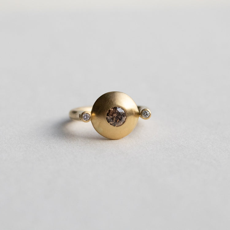 Modern 18ct yellow gold ring with a matte circular spinning disc speckle set with a central natural brown cognac diamond and white side diamonds, paper shot front