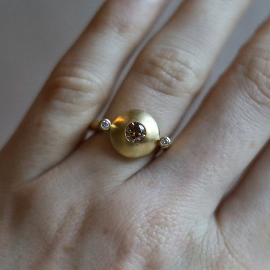 Modern 18ct yellow gold ring with a matte circular spinning disc set with a central natural brown cognac diamond and white side diamonds, worn on the hand.