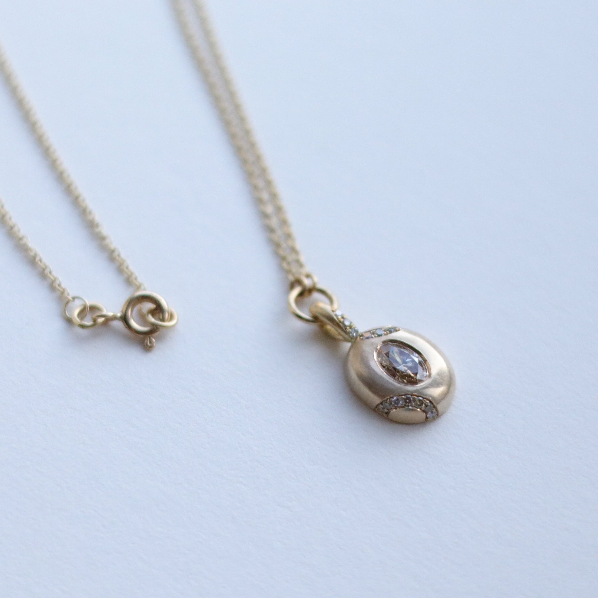 Goldfinch jewellery Close up of solid 9ct yellow gold oval pendant necklace with pave champagne diamond details and a flush set brown oval natural diamond. On a loop in-line on a fine chain. photographed on paper, view of the front, with clasp in shot.