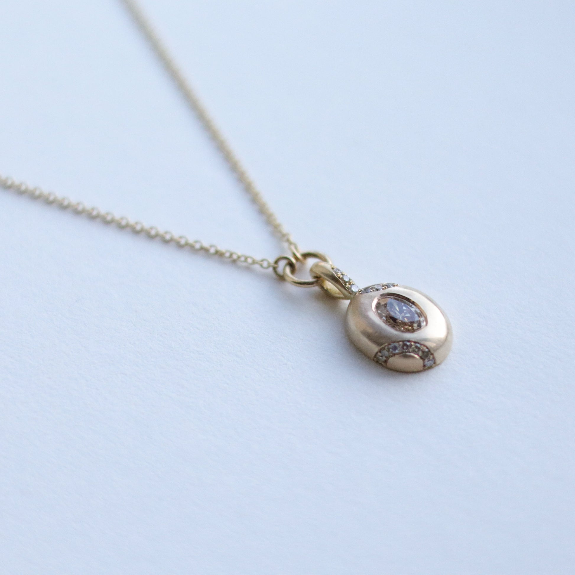 Close up of solid 9ct yellow gold oval pendant necklace with pave champagne diamond details and a flush set brown oval natural diamond. On a loop in-line on a fine chain. photographed on paper, view at angle.