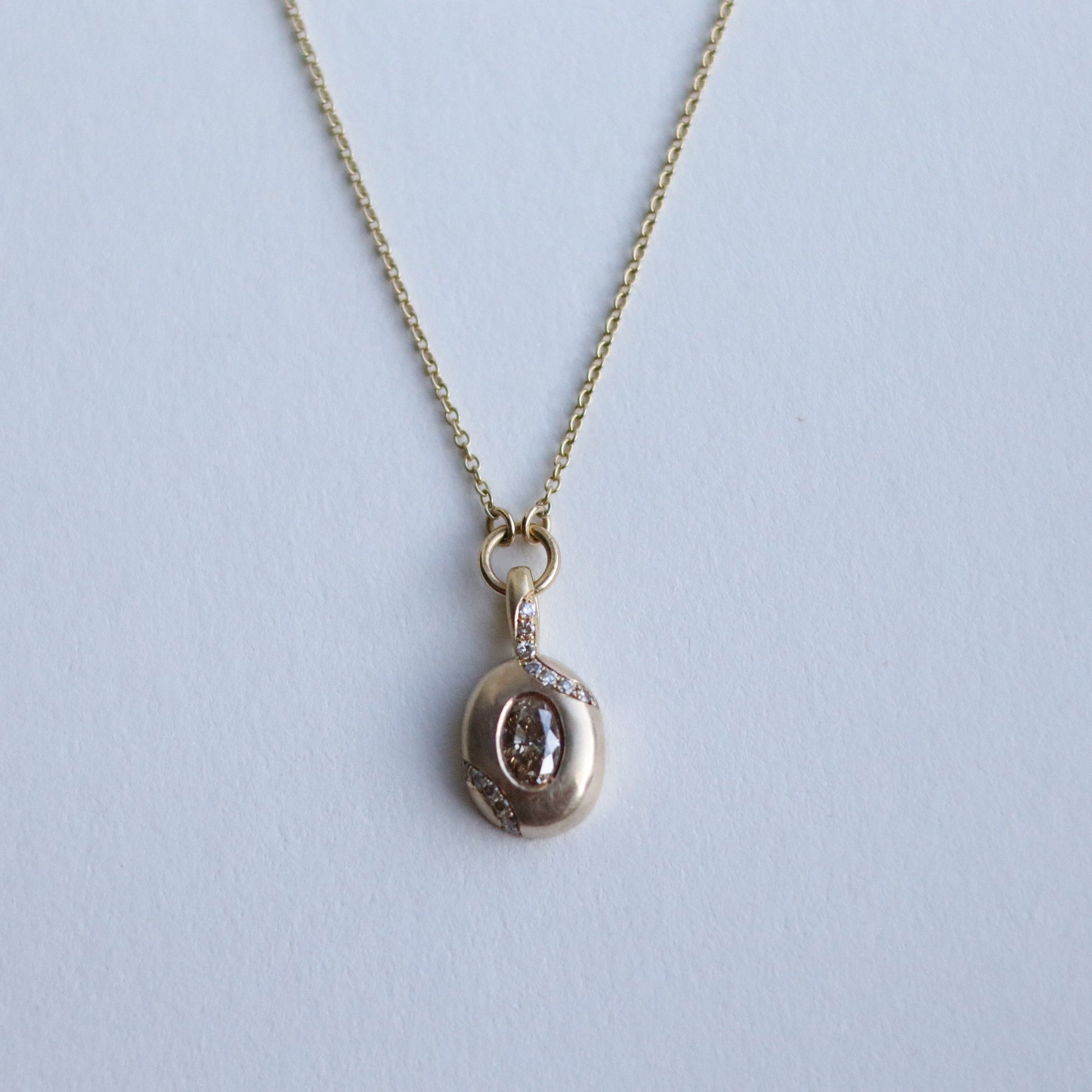 Close up of solid 9ct yellow gold oval pendant necklace with pave champagne diamond details and a flush set brown oval natural diamond. On a loop in-line on a fine chain. photographed on paper, view of the front