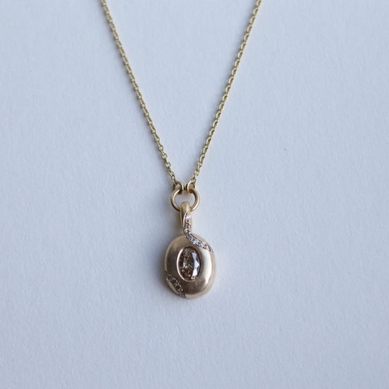Close up of solid 9ct yellow gold oval pendant necklace with pave champagne diamond details and a flush set brown oval natural diamond. On a loop in-line on a fine chain. photographed on paper, view of the front