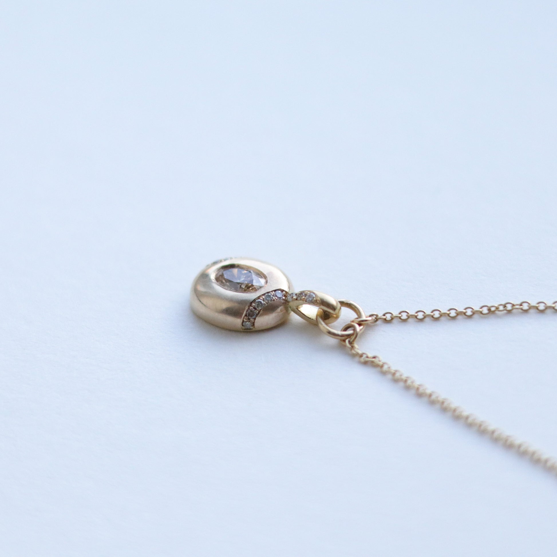 Close up of solid 9ct yellow gold oval pendant necklace with pave champagne diamond details and a flush set brown oval natural diamond. On a loop in-line on a fine chain. photographed on paper, view of the top
